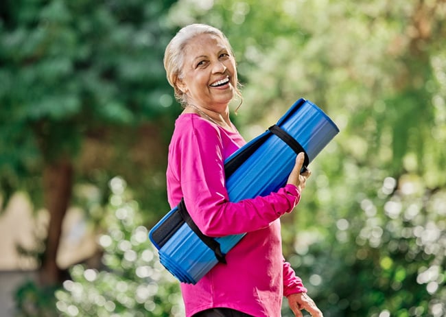 Senior woman outside smiling and holding a rolled-up yoga mat